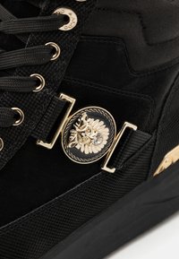 Svart sneaker med en blandning av mocka och texturväv, med guldhardware och en lejon-design som detaljer på en rund platta.