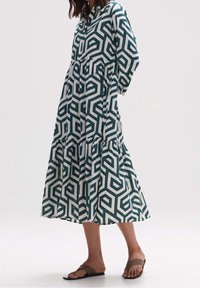 Geometriskt mönstrad klänning i teal och vitt, med långa ärmar, knäppning vid halsen och en skiktad kjoldesign. Tillverkad av lättviktsmaterial.