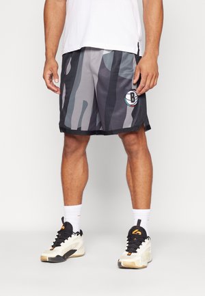 Hombre con pantalones cortos de baloncesto camuflaje negro y gris con el logo de Brooklyn Nets, calcetines blancos y zapatillas blancas y negras, posando contra un fondo blanco.