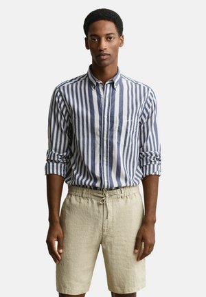 Homme portant une chemise à boutons à rayures verticales bleues et blanches avec les manches retroussées et un short beige à cordon, debout contre un fond uni.