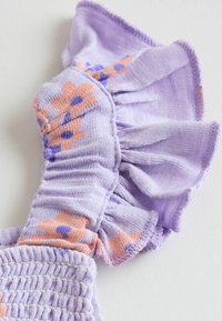 Gros plan sur un tissu lavande avec un imprimé floral orange et violet et un détail de bord volanté sur fond blanc.