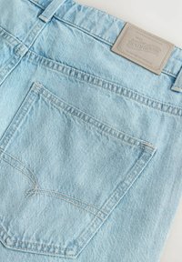 Jeans en denim bleu clair avec une poche arrière ornée de coutures blanches et une étiquette en cuir beige au niveau de la taille portant les détails de la marque.