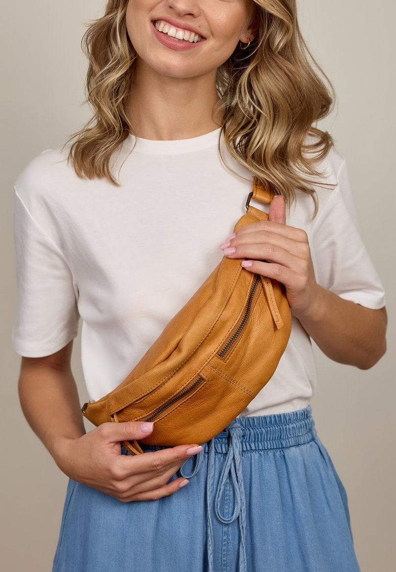 Sveća žena u bijelom majici i plavoj suknji drži bež kožnu torbicu s patentnim zatvaračem ispred bež pozadine.