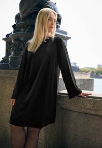 Schwarzes langärmliges Kleid mit lockerem Schnitt, weichem Stoff und leichtem Glanz. Hat einen runden Halsausschnitt, der mit dekorativen Akzenten verziert ist.