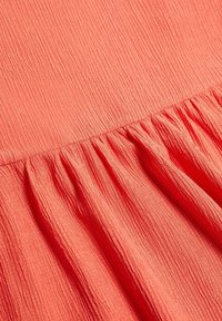 Tissu de couleur corail avec un motif texturé et froissé, présentant un détail de volants froncés le long du bord inférieur.