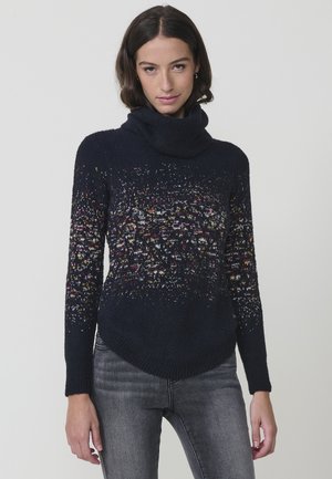 Suéter de cuello alto en color navy con un tejido texturizado, motas multicolores en la parte superior y un dobladillo curvado. Combinado con jeans grises.
