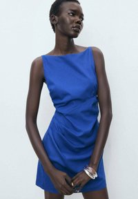 Robe sans manches bleue avec un col bateau, présentant un design froncé et une texture lisse. Bras orné d'un bracelet transparent.