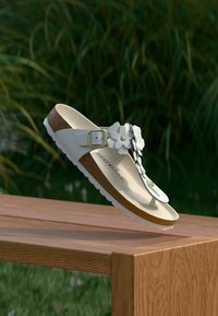 Λευκό σανδάλι Birkenstock με φελλό σόλας και ανθισμένες διακοσμήσεις που ακουμπά στην άκρη ενός ξύλινου παγκακιού σε εξωτερικό χώρο με φόντο πράσινη βλάστηση.