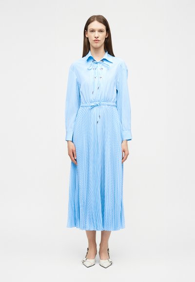 Γυναίκα όρθια, φορώντας ένα μακρυμάνικο midi φόρεμα σε ανοιχτό μπλε και λευκή ρίγα με πλισέ φούστα και λευκά μυτερά παπούτσια.