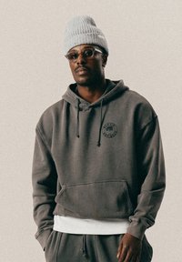 Mężczyzna w szarej bluzie z kapturem z logo drzewa, w dopasowanych spodniach, jasnoszarym dzianinowym beretcie, w okularach i małych kolczykach w kształcie kółek, stojący na jednolitym tle.