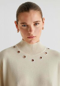 Cremefarvet strikket sweater med høj ribbede krave og sølvøje detaljer omkring halsudskæringen. Blød tekstur og en afslappet pasform.