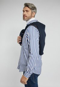 Granatowy hoodie narzucony na ramię, w połączeniu z niebieską i białą koszulą w paski z guzikami. Dżinsowe spodnie dopełniają całość.