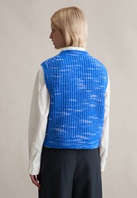 Nieprzemakalny niebieski kamizelka z teksturowanym wzorem, z okrągłym dekoltem. Noszona na białej koszuli z kołnierzykiem, ma luźny krój i brak zapięcia.