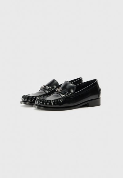 Schwarze Lackleder-Slipper mit runder Zehenpartie, dekorativer Steppnaht und einem kleinen Schnallenakzent. Mit einem niedrigen Blockabsatz.