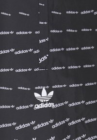Svart tyg med upprepade vita "adidas"-loggor och trefoil-grafik; slät textur, modern design, med en delvis dold söm.
