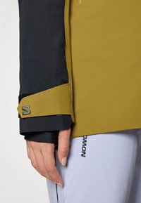 Voděodolná bunda se žlutou a černou tkaninou. Obsahuje suchý zip na manžetách, elegantní design a detail s logem na rukávu.