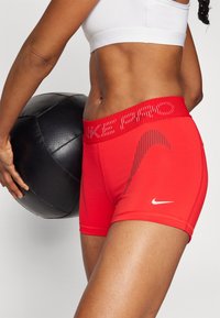 Crvene atletske kratke hlače s prilagodljivim dizajnom, koje imaju široku, strukturiranu elastiku oko struka i suptilne crne linije za detalje. Nike logo u bijeloj boji.