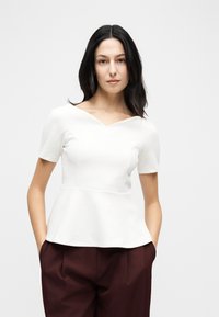 Femme aux cheveux foncés portant un haut péplum blanc à manches courtes et un pantalon marron foncé, debout les mains dans les poches, devant un fond uni.