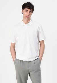 Witte polo met een klassieke kraag, korte mouwen en een subtiel logo. Gecombineerd met lichtgrijze broeken. Gladde stofstructuur.