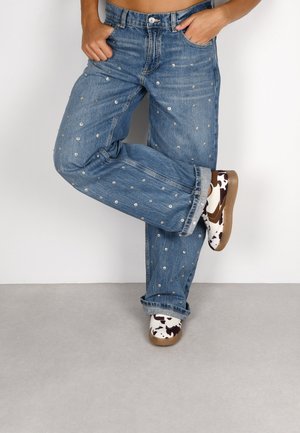 Jeans in denim blu con decorazioni argentate, vestibilità comoda e orli risvoltati, abbinati a scarpe bianche con stampa di mucca marrone e suole in gomma.