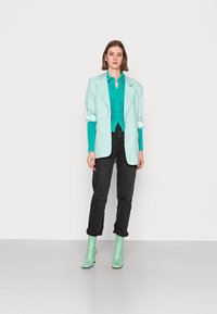 Blazer oversized verde menta sobre uma camisa azul ciano, combinado com jeans pretos e botas de tornozelo verde menta a condizer. Fundo simples.