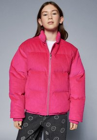 Roze baršunasta puf jakna s visokim ovratnikom, zatvaranjem na zip i džepovima s flasterima. Nosi se preko tamnih hlača s uzorkom nasmijanih lica.