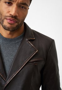 Braune Lederjacke mit Reverskragen und sichtbaren Steppnähten, geschichtet über einem grauen Strickpullover. Mit Reißverschlusstasche.