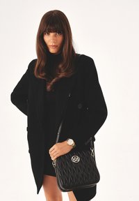 Borsa a spalla nera trapuntata con accessori in oro, caratterizzata da un logo prominente. Il modello indossa un cappotto nero e un orologio dorato. Texture liscia.