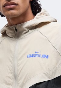 Крупен план на човек, носещ бежово яке с качулка, цип и син текст "BERLIN" и лого на Nike на гърдите.