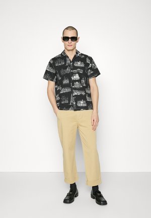 Mężczyzna w czarnych okularach przeciwsłonecznych, czarnej koszuli z krótkim rękawem z białymi architektonicznymi nadrukami, beżowych spodni 7/8, czarnych skarpetach i czarnych mokasynach.