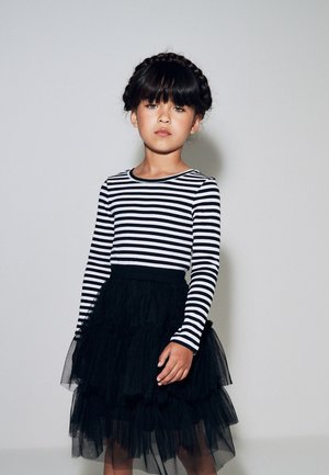Giovane ragazza con i capelli intrecciati indossa una maglia a maniche lunghe a righe bianche e nere e una gonna nera a strati in tulle, in piedi davanti a uno sfondo semplice.