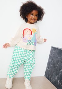 Sudadera crema con gráficos coloridos, combinada con pantalones verdes de cuadros. Presenta mangas largas y un ajuste relajado, complementado con zapatillas blancas.