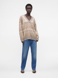 Personne debout face à l'avant, portant un pull oversize beige à motif zigzag, un jean bleu ample et des talons ouverts, sur un fond uni.