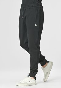 Pantaloni sportivi neri realizzati in tessuto morbido, dotati di una cintura elastica, tasche laterali e un piccolo logo ricamato sulla coscia. Abbinati a sneakers bianche.