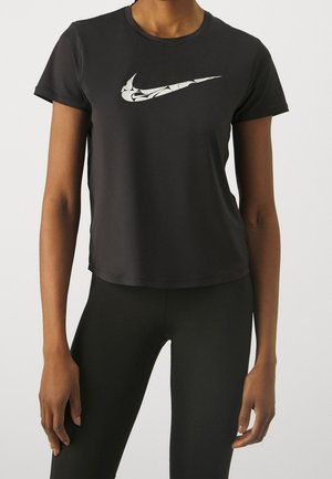Personne portant un t-shirt noir moulant Nike avec un logo swoosh blanc à motif et un legging noir, debout devant un fond clair uni.