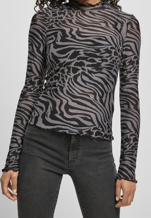 Vrouw in een lange, aansluitende top met een grijs en zwart zebra patroon en donkergrijze hoge taille jeans, met ontspannen handen langs de zijkanten.