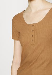 Brun ribbet kortærmet top med rund halsudskæring og fem knaplukninger. Stoffet har en glat, strækbar tekstur med et tætsiddende design.