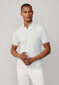 Šedé polo tričko s klasickým límcem a krátkými rukávy, s jemnou texturou. Model má na sobě bílé kalhoty, ruce sepnuté vpředu.