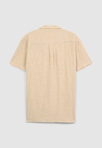 Camicia beige a maniche corte realizzata in tessuto leggero. Presenta un colletto a punta e una spalla posteriore con una piega. La texture appare leggermente strutturata.