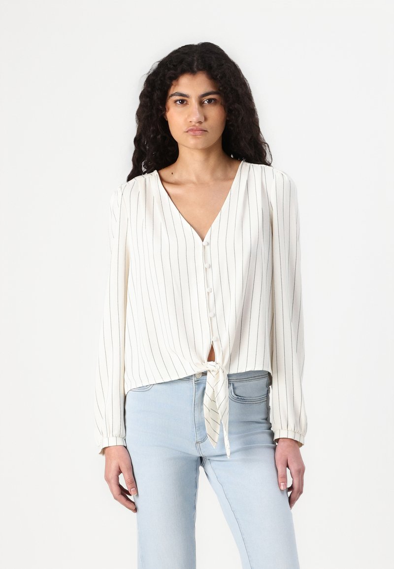White long-sleeve blouse with black vertical stripes, V-neck, button-down front, and tied hem at the waist. Soft fabric with a slight sheen.