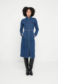 Modra jeans srajčna obleka z dolgimi rokavi, zapiranjem na gumbe, dvema prsnikoma in pasom, z razporkom na spodnjem robu. Kombinirana s črnimi škornji do kolen.