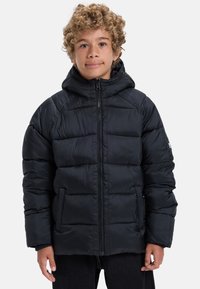 Pojke med lockigt hår som bär en svart puffig jacka med huva, står mot en enfärgad ljus bakgrund, vänd framåt.