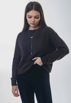 Ciemnobrązowy, dziergany sweter o luźnym kroju i długich rękawach. Noszony na czarnych spodniach, zestawiony z naszyjnikiem z srebrnym akcentem.