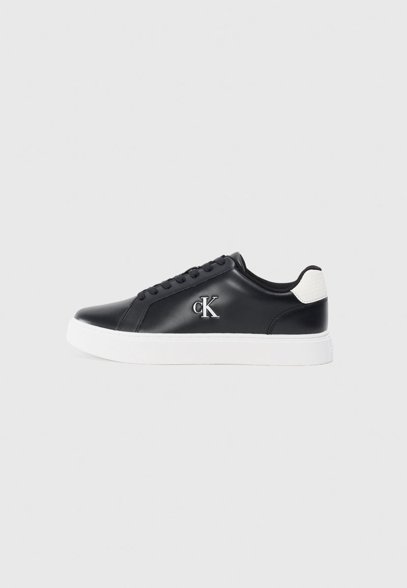 Tenis negri din piele cu talpă albă, vârf rotund, cinci găuri pentru șireturi și un logo argintiu "cK" pe partea exterioară. Accente texturate pe călcâiul alb.