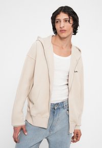 Beige rebrovaná mikina na zips s prednými vreckami a kapucňou na šnúrku, nosená cez biely top s tenkými ramienkami, doplnená svetlými džínsami.