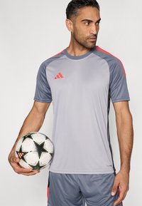 Γκρι αθλητική φανέλα με κόκκινες λεπτομέρειες, κοντά μανίκια και στρογγυλή λαιμόκοψη. Το λογότυπο της Adidas εμφανίζεται στο μπροστινό μέρος. Ο παίκτης κρατάει μια ασπρόμαυρη ποδοσφαιρική μπάλα.