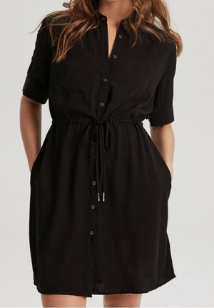 Femme portant une robe noire à manches courtes avec des boutons sur le devant, une taille cintrée par un cordon et des poches, debout devant un fond clair uni.