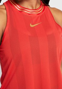 Czerwona sportowa koszulka bez rękawów z siateczkową strukturą, złotymi akcentami i trzema poziomymi złotymi paskami wokół dekoltu. Logo Nike widoczne.