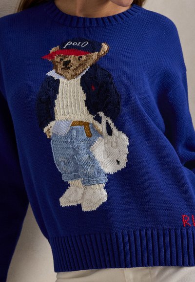 Μπλε πλεκτό πουλόβερ με γραφικό αρκούδας σε casual outfit, με κόκκινο καπέλο, λευκό πουλόβερ και τζιν, και μια λευκή τσάντα. Κόκκινο κείμενο στο τελείωμα.