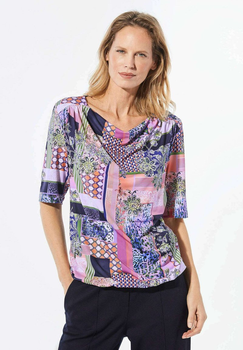 Blouse multicolore en tissu léger, avec un col drapé, des manches courtes et un motif mixte de fleurs, de formes géométriques et de rayures.